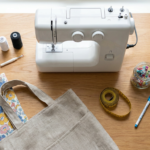 Flat lay of core beginner sewing tools, including a sewing machine, scissors, neutral threads, pins, measuring tape, seam ripper, marking tool, iron, and colorful cotton fabric beside a simple sewing project.